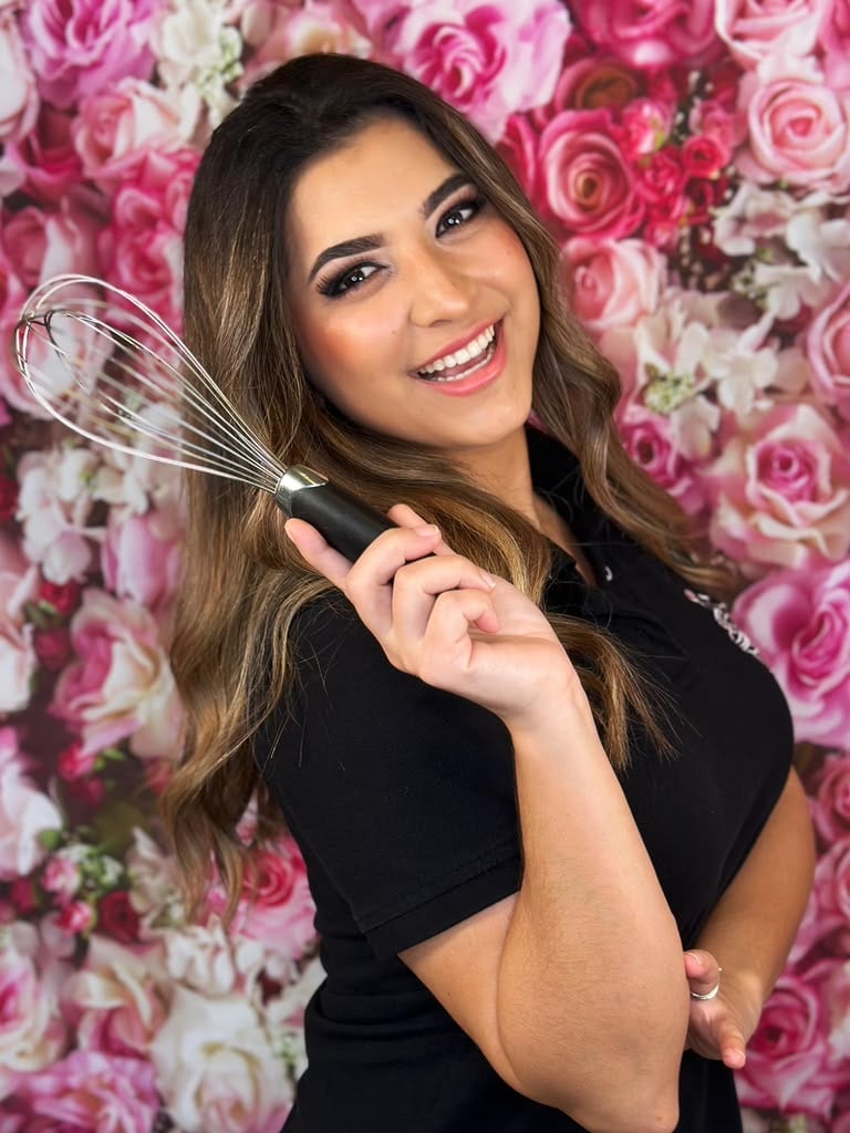 Woman holding a whisk and smiling in front of a pink and white rose flower backdrop
