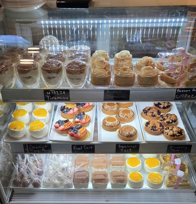 Bakery display case with tiered shelves showing cakes, pastries, cookies, and desserts with price labels and LED lighting