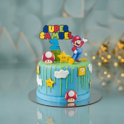 Super Mario themed birthday cake with colorful letter topper, character figurines, red and yellow mushrooms, and stars on light blue frosting