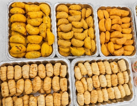 Five aluminum trays of assorted fried pastries and savory snacks, including golden donut-like pastries and elongated fried dough pieces