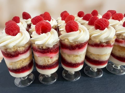 Row of layered dessert cups with graham cracker base, raspberry filling, whipped cream, and fresh raspberries on top