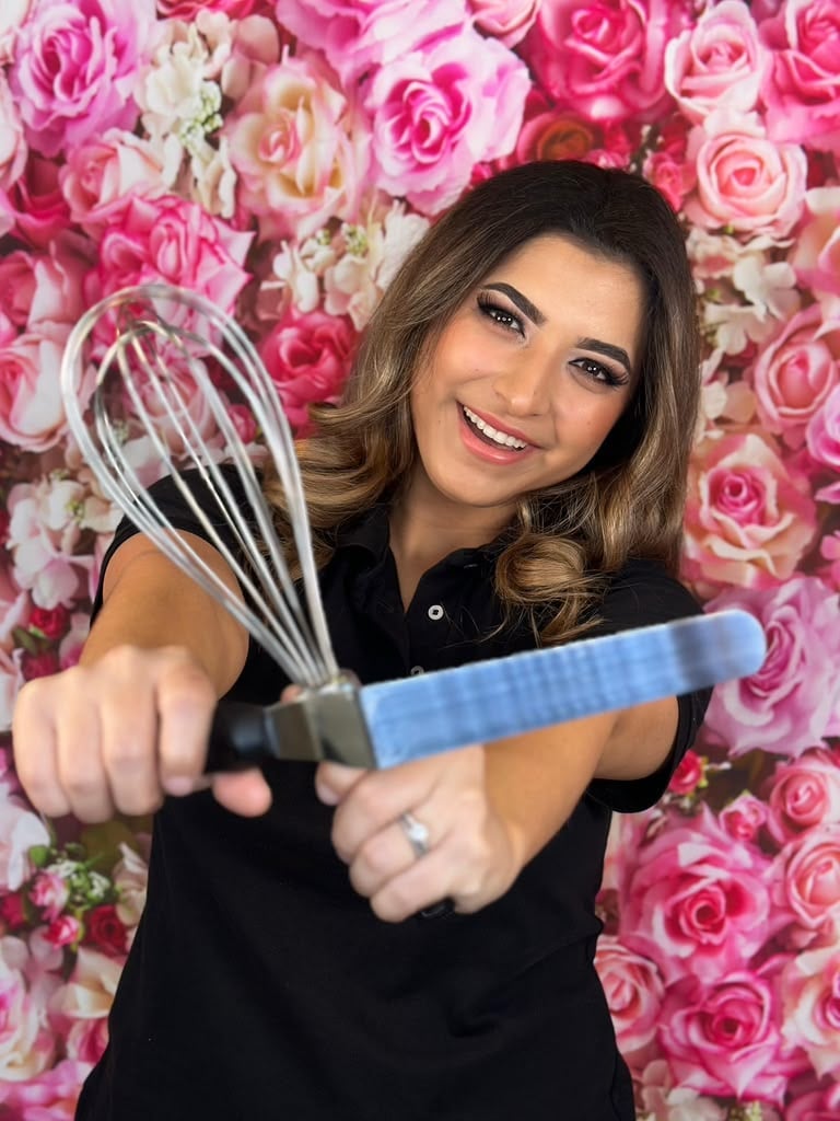 Smiling woman holding kitchen utensils in front of a pink rose flower backdrop